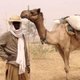 Man in head scarf and sun hat standing by camel, with an arid backdrop (Yobe river basin)