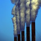 The tops of five chimney stacks releasing thick fumes, set against the blue sky.